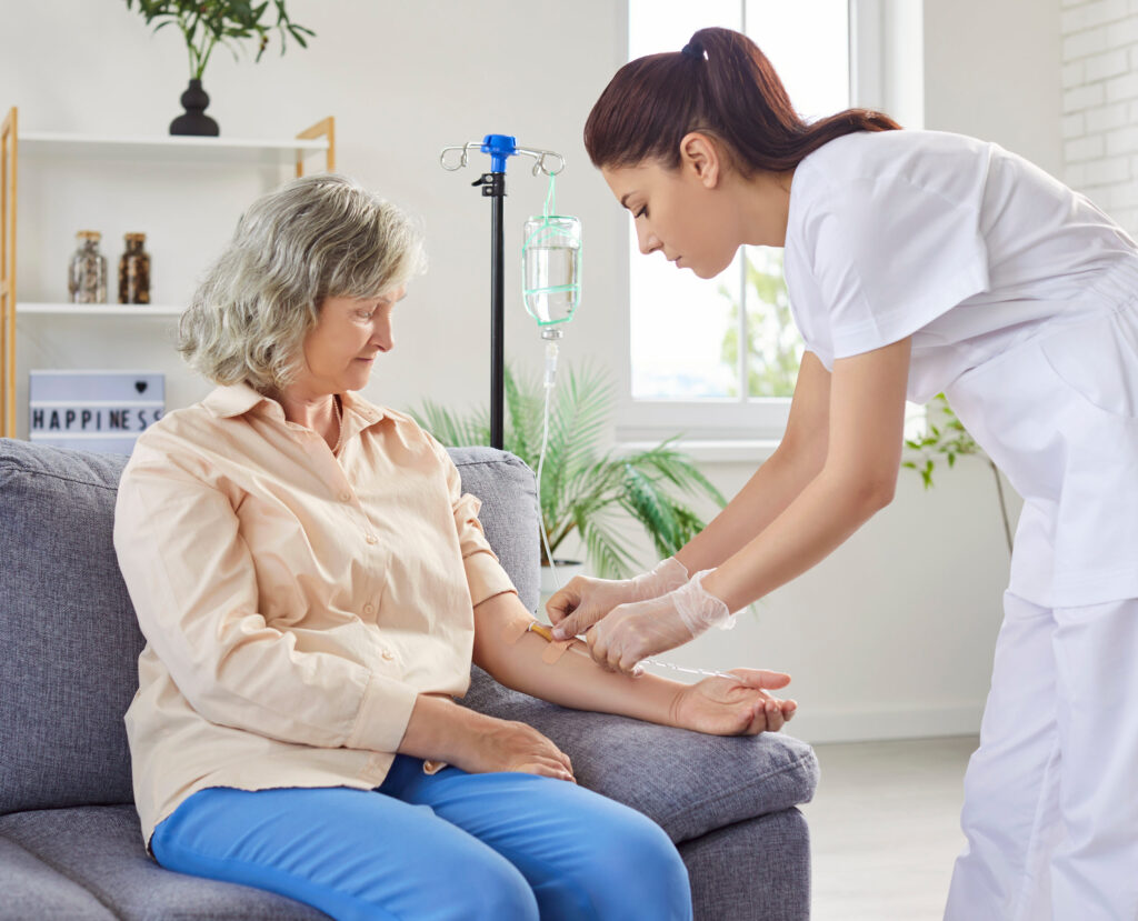 A senior woman sitting on the couch in her living room receiving a vitamin IV drip from an experienced nurse.